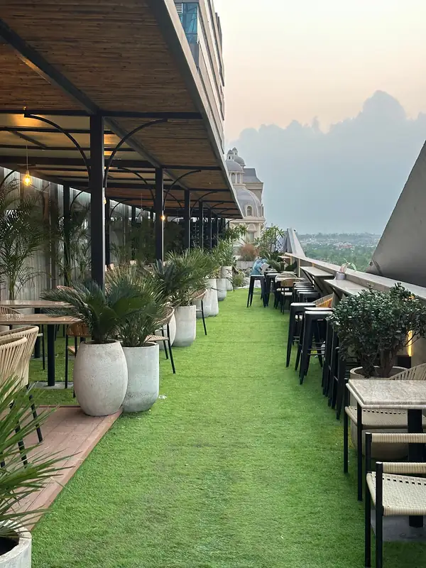 The rooftop terrace in the late afternoon, with a bamboo pergola, palms in white planters, bar stools along the edge and the city beyond.