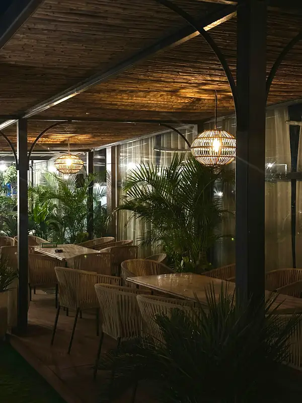 The rooftop terrace at night, lit by woven pendant lamps over wicker chairs and palms.