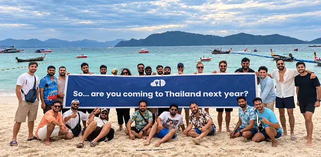 The team on a beach in Phuket in 2025, holding a Next Order banner that reads: So... are you coming to Thailand next year?